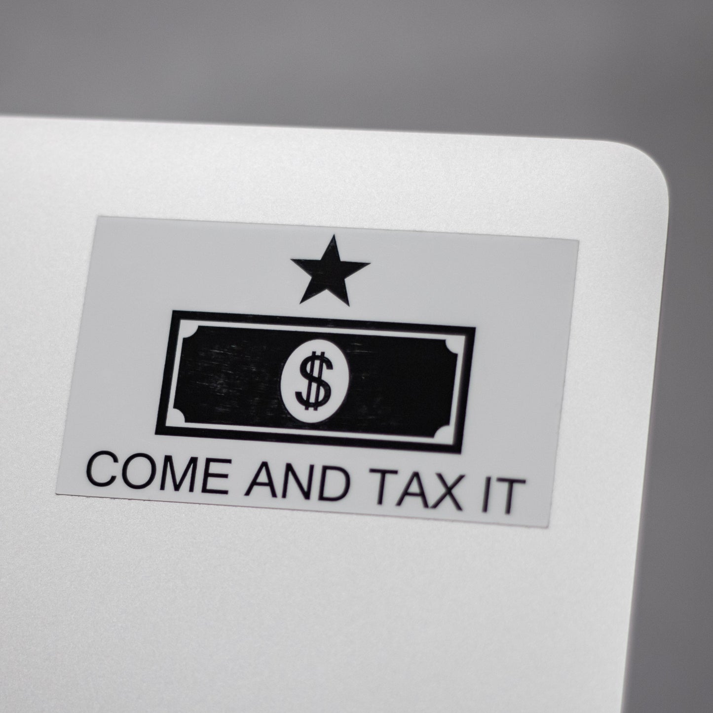 Sticker with a dollar bill design and text 'COME AND TAX IT' on the back of a metallic laptop.