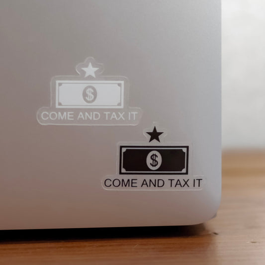 Close-up of a metallic laptop back with two small 'COME AND TAX IT' stickers. Laptop is on a wooden surface.