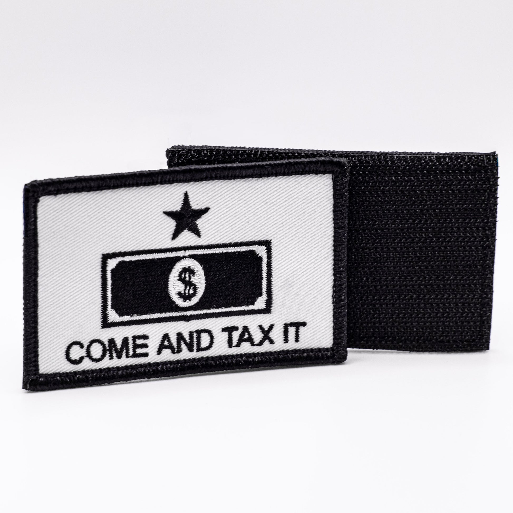 A white patch featuring in black the words "come and tax it" under a outline of a dollar bill, with a star above both. The hook and loop backing of another patch is shown in the background