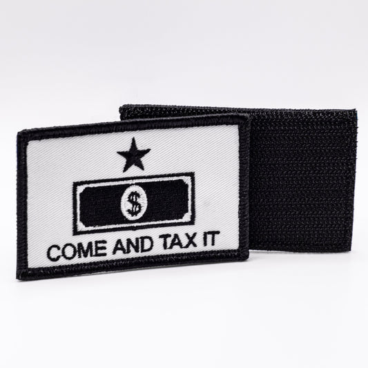 A white patch featuring in black the words "come and tax it" under a outline of a dollar bill, with a star above both. The hook and loop backing of another patch is shown in the background