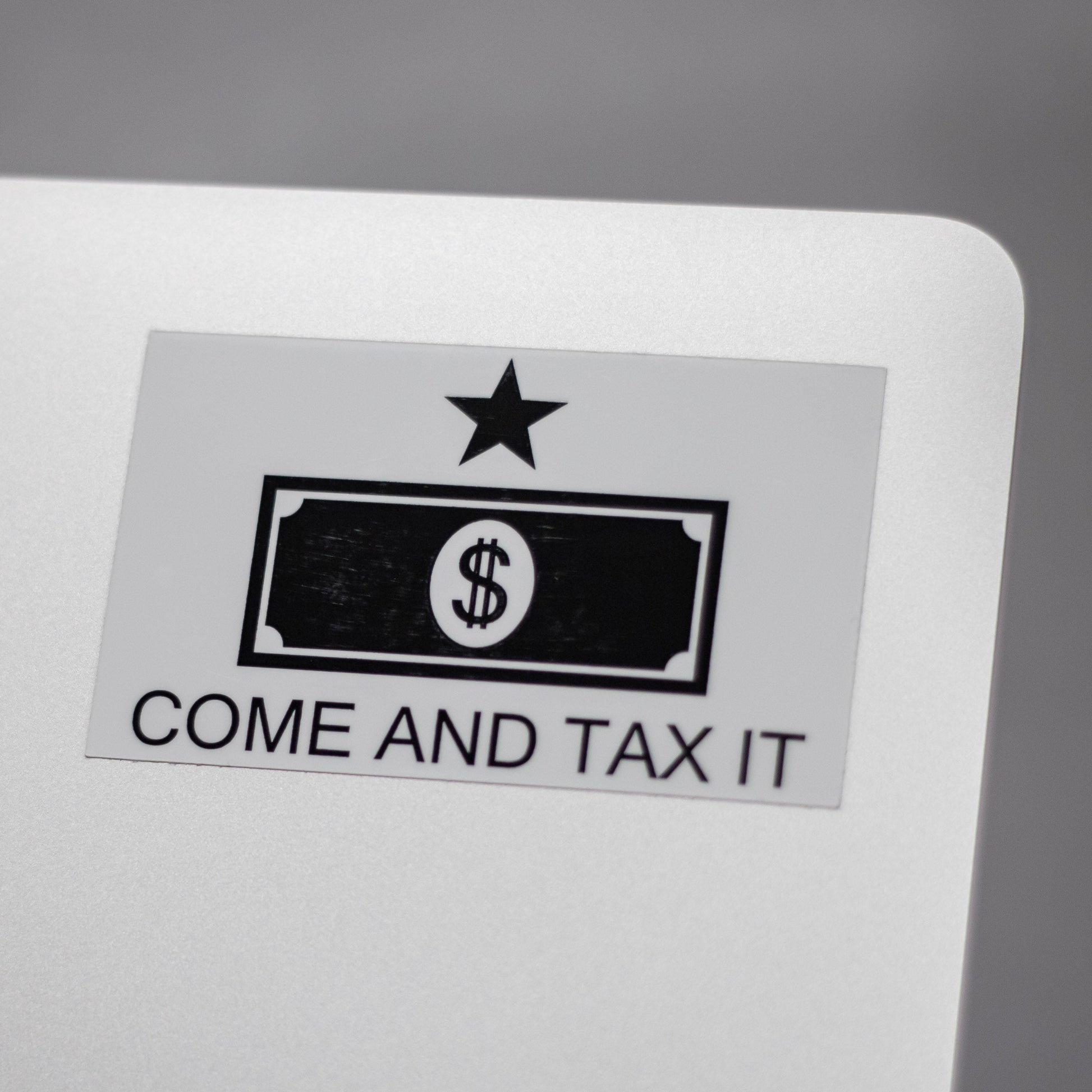 Sticker with a dollar bill design and text 'COME AND TAX IT' on the back of a metallic laptop.