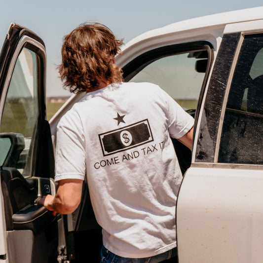 A photo of a man getting into a white pickup truck while wearing a white shirt which on the back says "COME AND TAX IT" beneath a stylized dollar bill and star.